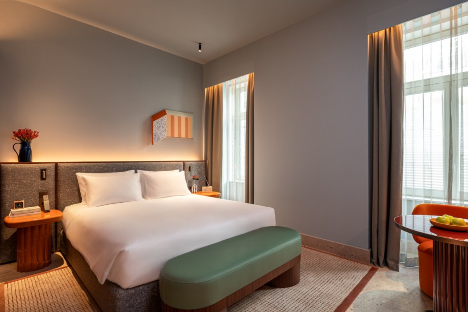 A modern hotel room featuring a large bed with white linens, contemporary decor, a small seating area with a round table, and large windows with sheer curtains.