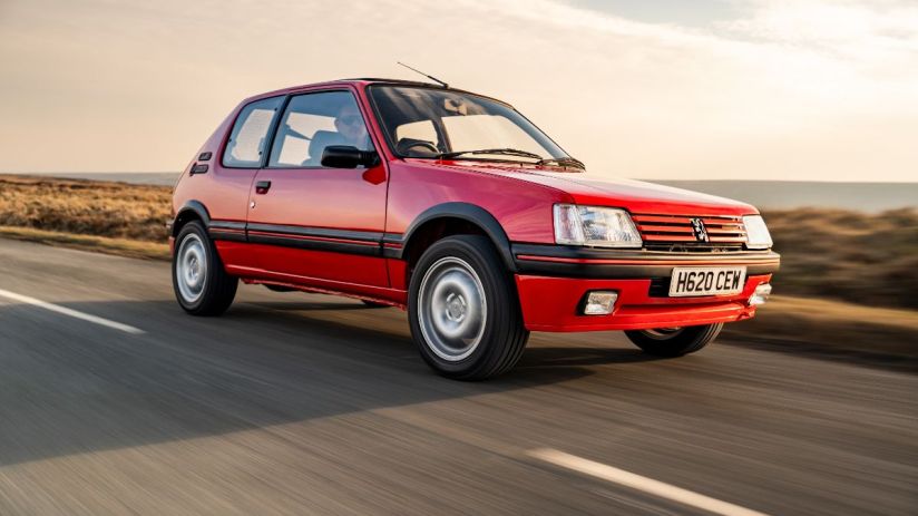 Vintage Peugeot 205 driving on a scenic road, showcasing classic design and compact size for a news feature on iconic cars