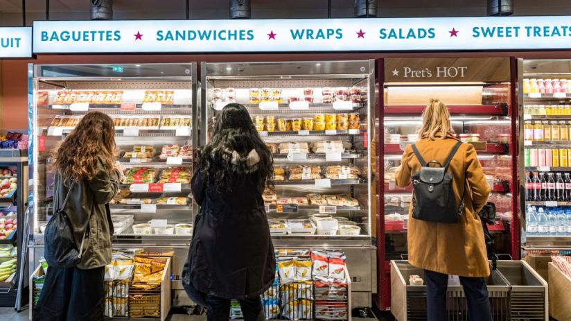 A busy Pret A Manger storefront with customers entering and exiting during lunchtime in a bustling city center.