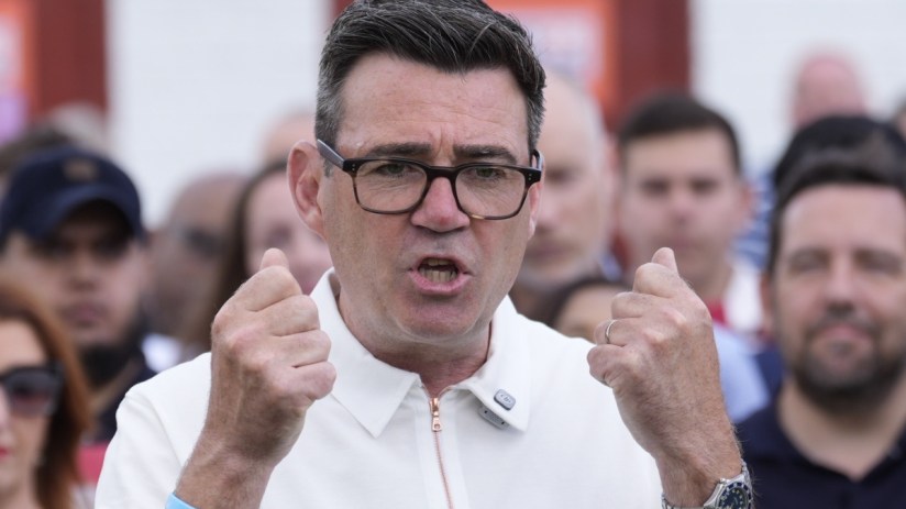 Andy Burnham speaking at a press conference, addressing current issues, wearing a suit and tie, with a serious expression.