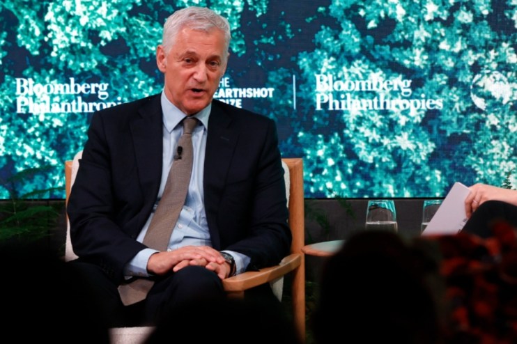 Standard Chartered CEO Bill Winters at an event, wearing a suit, speaking into a microphone against a corporate backdrop.