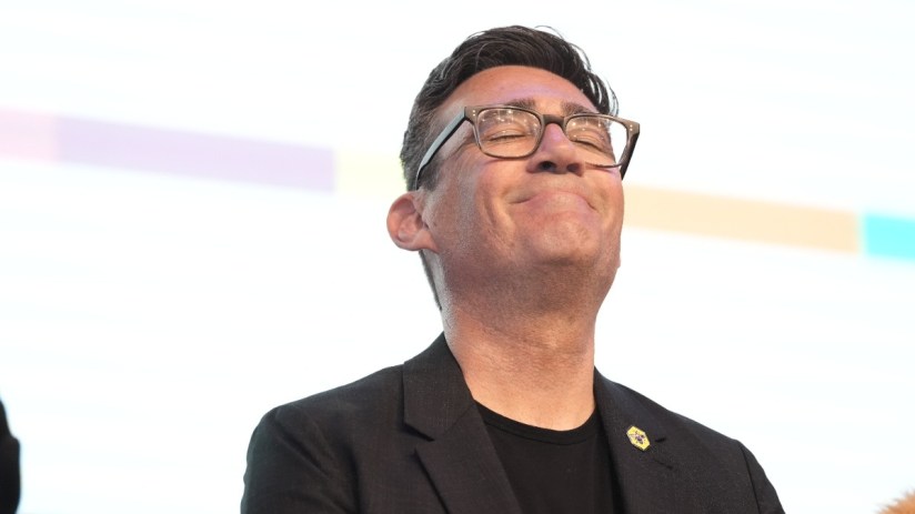 Andy Burnham smiling at a public event, wearing a suit and tie, representing positive leadership and community engagement.