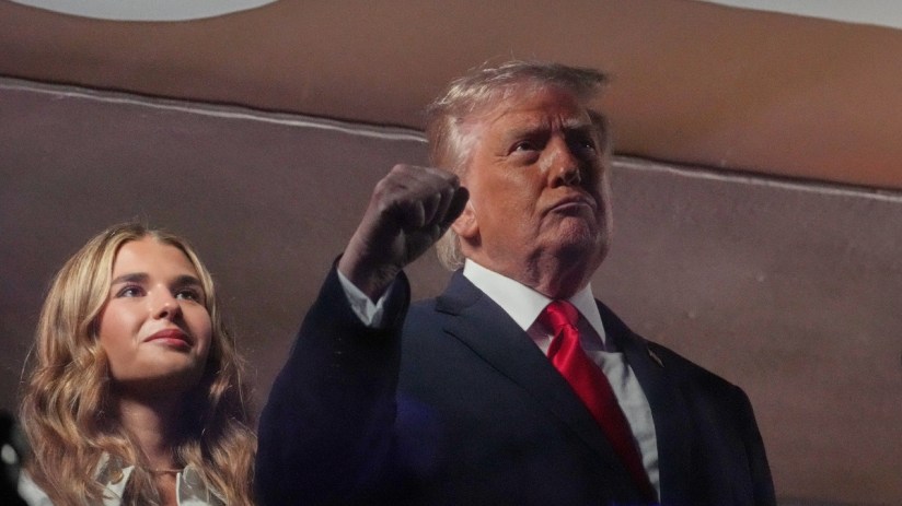 Donald Trump addressing media at a press event, wearing a suit and tie, with reporters and cameras in the background.