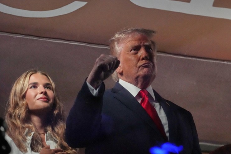 Donald Trump addressing media at a press event, wearing a suit and tie, with reporters and cameras in the background.