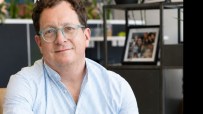 Smiling man with light blue glasses and striped shirt in an office setting.
