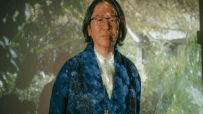 Person with long dark hair and glasses wearing a blue patterned jacket, projection of greenery behind them.