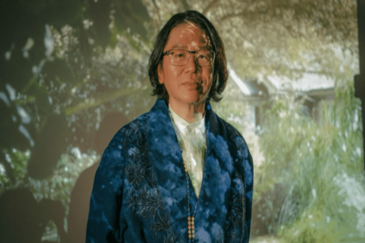 Person with long dark hair and glasses wearing a blue patterned jacket, projection of greenery behind them.