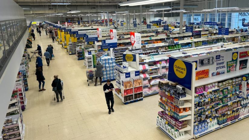 Overhead view of a brightly lit Tesco store interior with shoppers, product aisles, and Clubcard Prices signage.