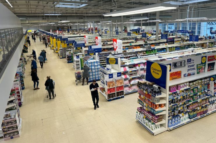 Overhead view of a brightly lit Tesco store interior with shoppers, product aisles, and Clubcard Prices signage.