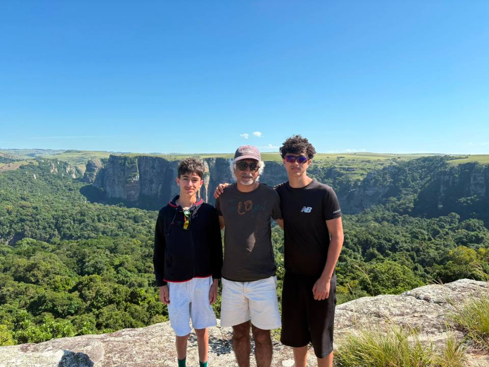 Three people standing on a rocky cliff overlooking a vast green canyon under a clear blue sky