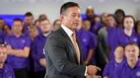 Alex Baldock in a suit and orange tie speaking to a crowd of people in purple shirts.