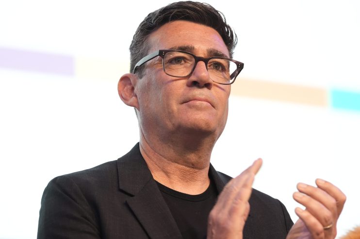Andy Burnham speaking at a press conference, wearing a suit, addressing key issues in Greater Manchesters development.