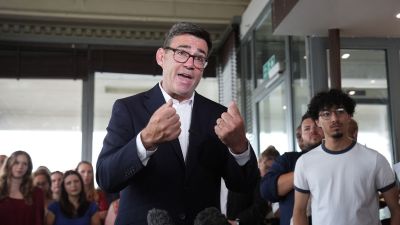 Andy Burnham speaking at a public event, emphasizing local governance and policy changes, wearing a suit and gesturing pas...