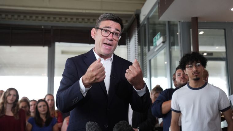 Andy Burnham speaking at a public event, emphasizing local governance and policy changes, wearing a suit and gesturing pas...