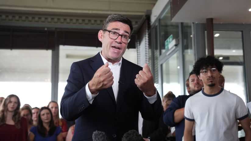 Andy Burnham speaking at a public event, emphasizing local governance and policy changes, wearing a suit and gesturing pas...