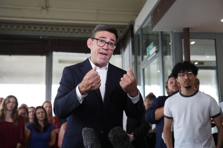 Andy Burnham speaking at a public event, emphasizing local governance and policy changes, wearing a suit and gesturing pas...