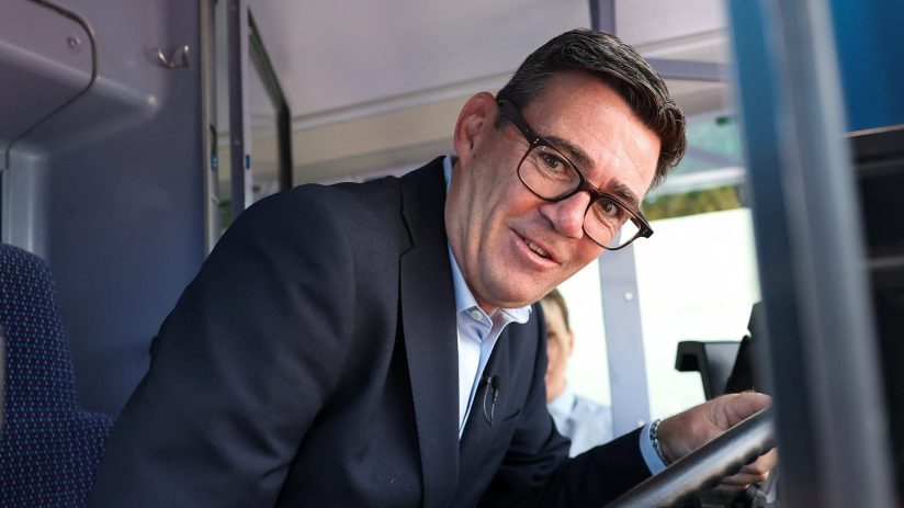 Andy Burnham smiling in a bus drivers seat, wearing glasses and a suit, addressing transport pledges.