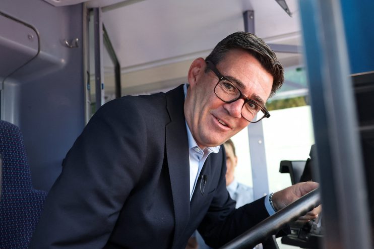 Andy Burnham smiling in a bus drivers seat, wearing glasses and a suit, addressing transport pledges.