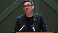 Andy Burnham speaking at a press conference, addressing current events and regional developments, wearing a suit and tie.