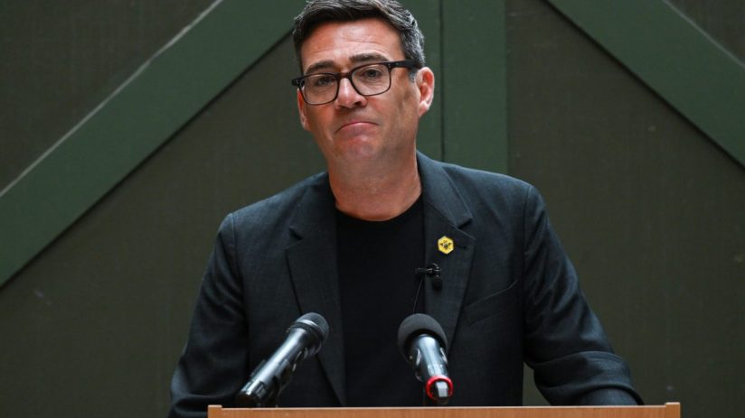 Andy Burnham speaking at a press conference, addressing current events and regional developments, wearing a suit and tie.