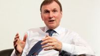 Archie Norman, business leader, speaking at a corporate event wearing a suit and tie, engaging with the audience.