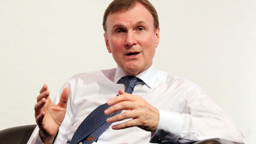 Archie Norman, business leader, speaking at a corporate event wearing a suit and tie, engaging with the audience.