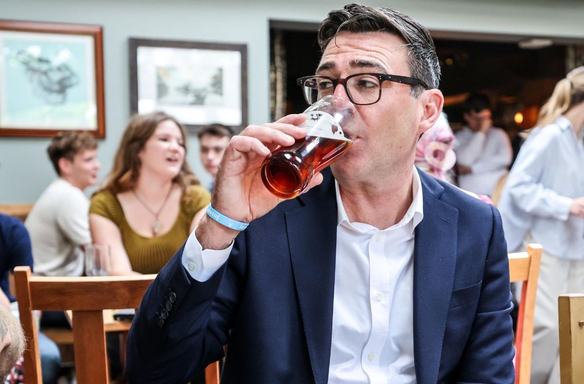 Andy Burnham, Mayor of Greater Manchester, drinks a pint of ale in a pub with people blurred in the background