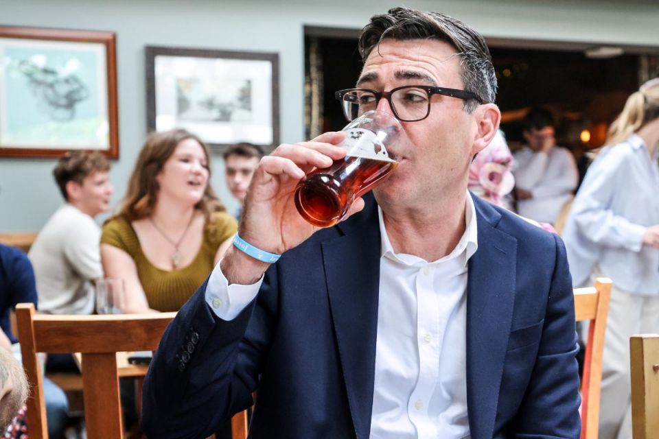 Andy Burnham, Mayor of Greater Manchester, drinks a pint of ale in a pub with people blurred in the background