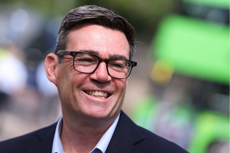 Andy Burnham, Mayor of Greater Manchester, smiling in glasses, dark jacket, light blue shirt, blurred green background.