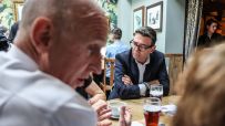 Andy Burnham, Mayor of Greater Manchester, in a dark suit and glasses, listening intently at a wooden table.