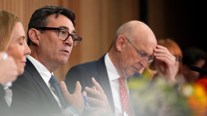 Andy Burnham, wearing glasses and a blue tie, speaks at a conference with a bald man in a red tie beside him.