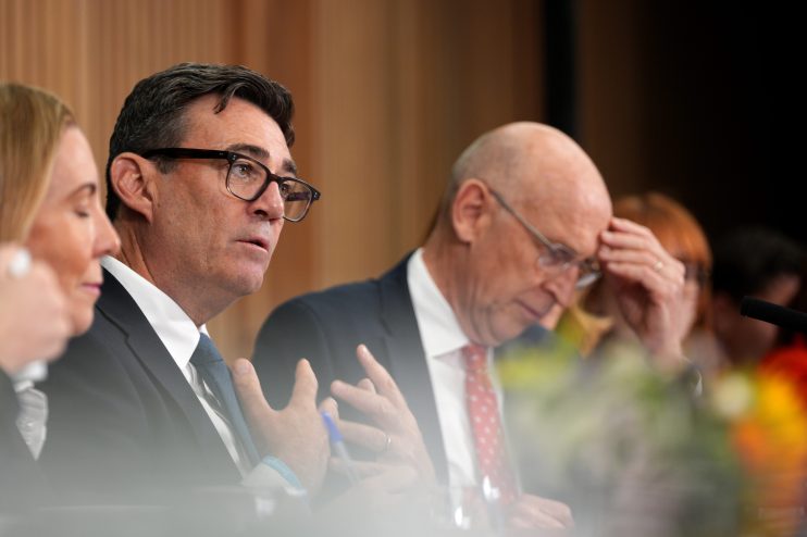 Andy Burnham, wearing glasses and a blue tie, speaks at a conference with a bald man in a red tie beside him.