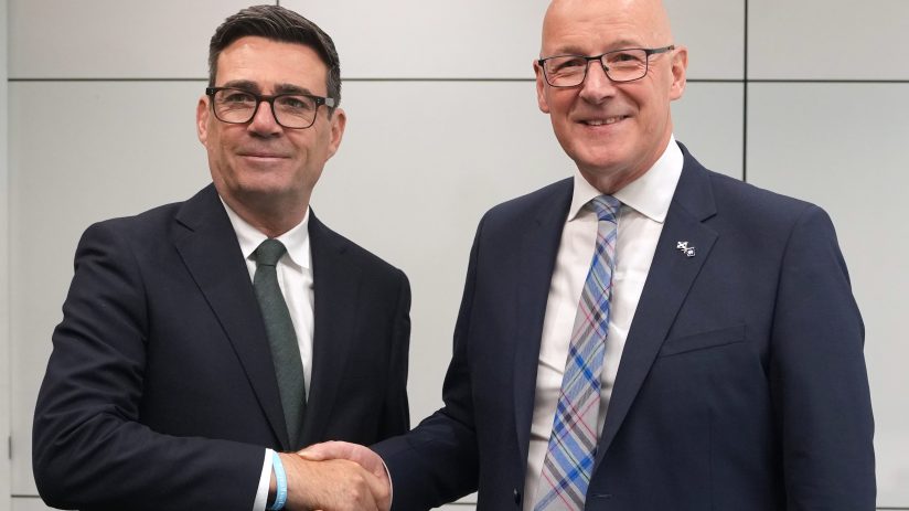 Andy Burnham and John Swinney shaking hands, both wearing dark suits and ties, in a professional setting.