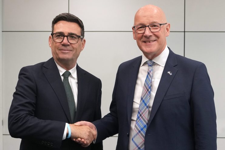 Andy Burnham and John Swinney shaking hands, both wearing dark suits and ties, in a professional setting.