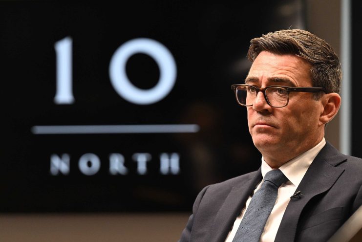 Andy Burnham, Mayor of Greater Manchester, in a suit and glasses, with a 10 NORTH logo in the background