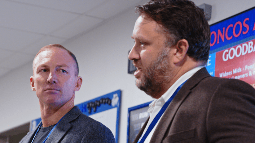 Two businessmen, one with a beard, in a meeting room with a red and blue digital display in the background.
