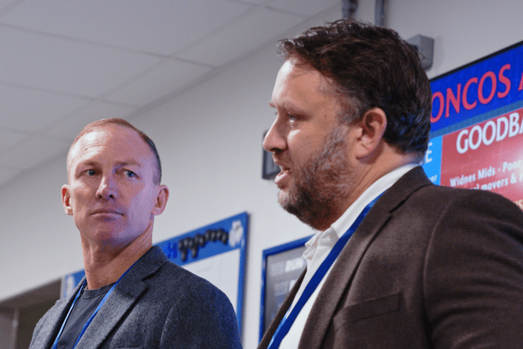Two businessmen, one with a beard, in a meeting room with a red and blue digital display in the background.