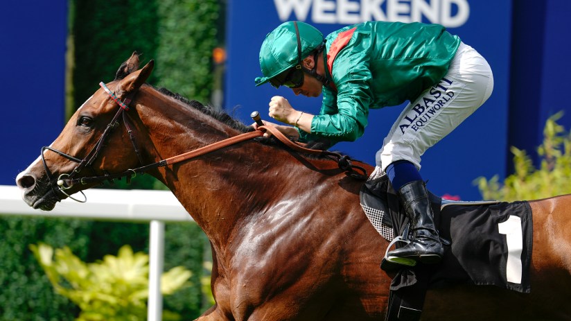 Jockey Calandagan on a brown racehorse, wearing green and white silks, racing past a blue sign with RACING WEEKEND