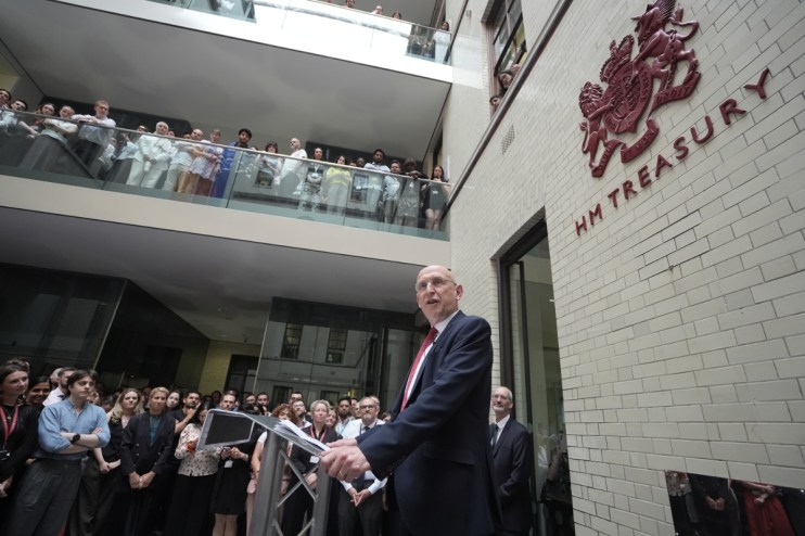 Chancellor Healey speaking at a podium before a crowd, with the HM Treasury sign visible on the brick building.