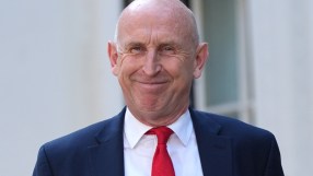 Chancellor John Healey smiling, wearing a navy suit, white shirt, and red tie.