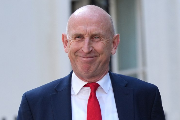 Chancellor John Healey smiling, wearing a navy suit, white shirt, and red tie.