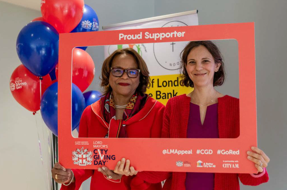 Two women smiling and holding a red City Giving Day frame, with red and blue balloons nearby.