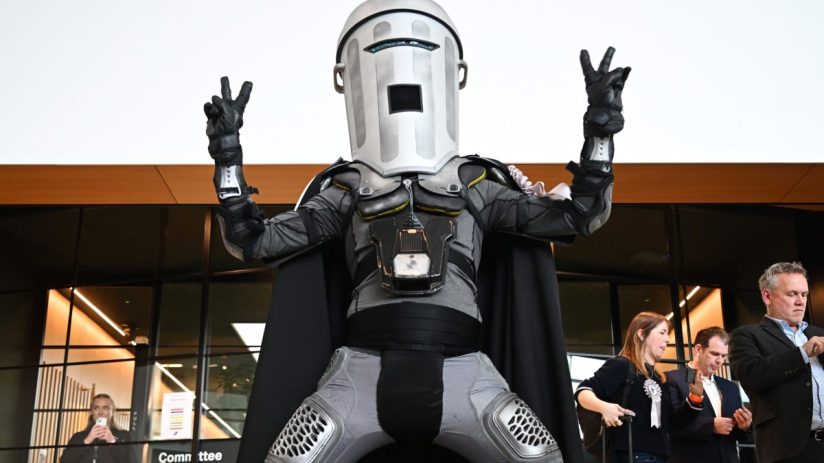 Count Binface wearing a silver mask and cape, standing in front of a podium during a press conference.