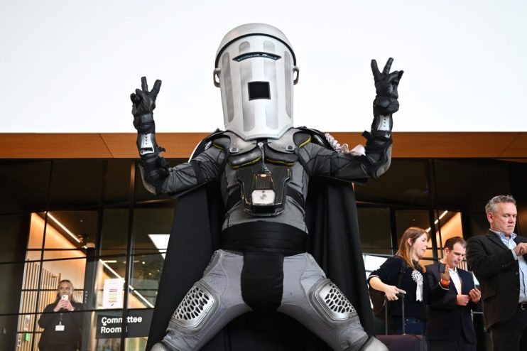 Count Binface wearing a silver mask and cape, standing in front of a podium during a press conference.