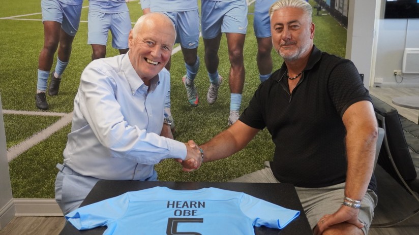 Barry Hearn and a man shaking hands over a light blue football jersey with HEARN OBE 5 printed on it.