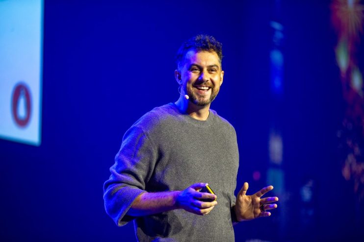 Daniel Hulme speaking at a business conference, wearing a suit, with a projector screen behind him displaying data graphs.