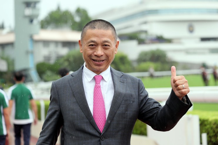 Danny Shum smiling at a press conference, wearing a suit, with a backdrop featuring sponsor logos and microphones on a table.