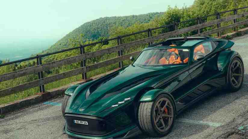 Dark green Donkervoort sportscar with exposed front wheels and orange interior, parked on a winding road with mountains