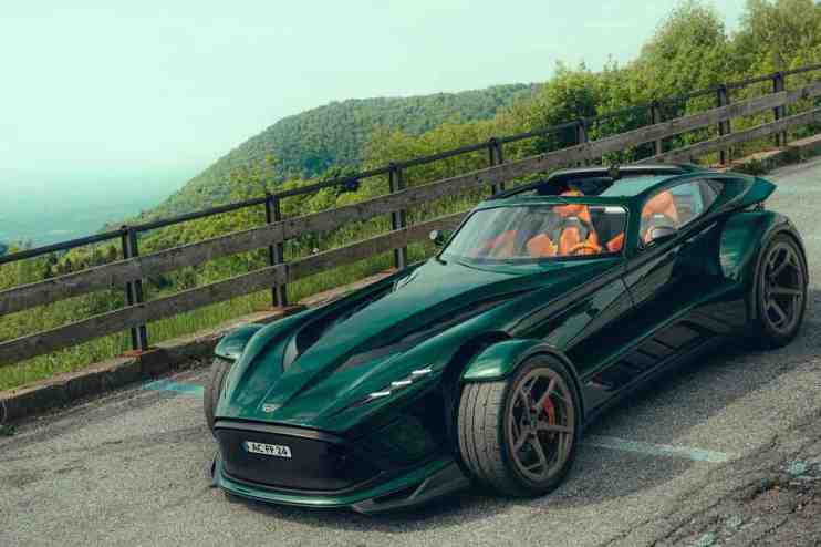 Dark green Donkervoort sportscar with exposed front wheels and orange interior, parked on a winding road with mountains