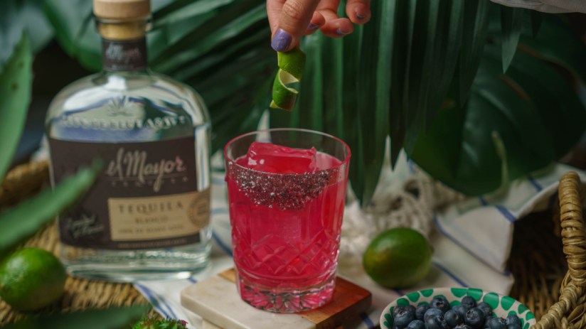 Hand garnishing a vibrant blueberry margarita with lime peel, alongside El Mayor tequila, limes, and blueberries.